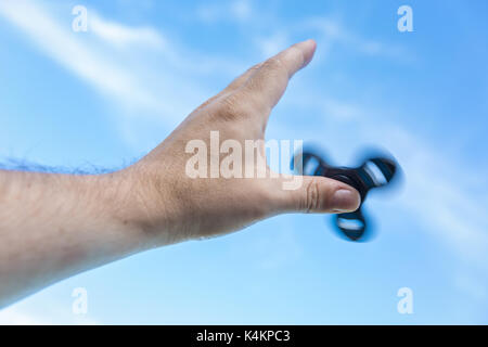 Man's hand holding a spinning fidget spinner dans sa main, le faisant tourner sur son pouce, contre le ciel bleu Banque D'Images