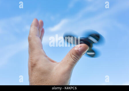 Man's hand holding a spinning fidget spinner dans sa main, le faisant tourner sur son pouce, contre le ciel bleu Banque D'Images