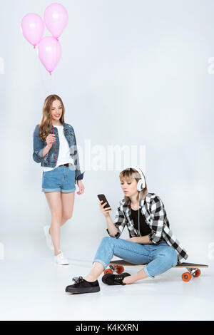 Jeune femme ballons roses et à la petite amie à l'aide d'écouteurs en tout en restant assis sur smartphone skateboard Banque D'Images