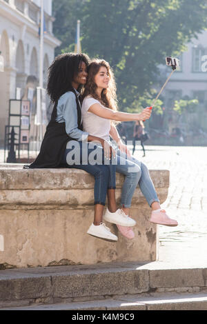 Sur toute la longueur de la magnifique petite amie assis sur la fontaine et prendre des autoportraits à l'aide de bâton selfies. Banque D'Images