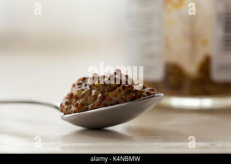 Comblé d'une cuillère à café de moutarde en grains entiers avec le pot de moutarde en verre faisant partie de l'arrière-plan flou. Tourné en lumière naturelle. Banque D'Images