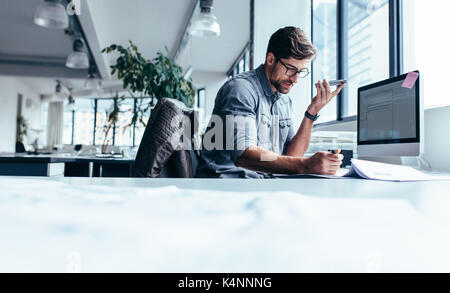 Businessman holding smart phone and working in office. Young caucasian man talking on téléphone à haut-parleur à son bureau. Banque D'Images