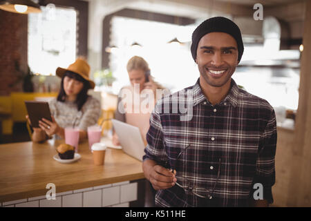 Portrait of smiling young man holding eyeglasses standing against female friends sitting at table in coffee shop