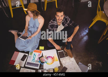 High angle portrait of smiling young male designer assis par collègue avec des feuilles sur le plancher du coffee shop Banque D'Images