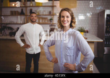 Portrait of smiling young woman with hand on hip contre l'ami du coffee shop Banque D'Images