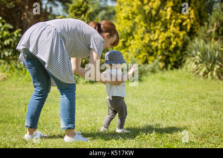 Petite fille de 14 mois fait ses premiers pas, à marcher sur ses propres sur la pelouse lors d'une journée ensoleillée ; aider sa mère en le tenant par derrière Banque D'Images