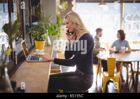 Side view of young blonde Woman talking on mobile phone while using laptop at cafe Banque D'Images