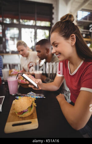 Vue latérale du smiling young woman text messaging while sitting avec burger at table in coffee shop Banque D'Images