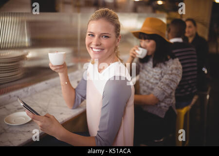 Portrait of smiling young woman drinking coffee lors de l'utilisation de téléphone mobile à l'encontre in coffee shop Banque D'Images