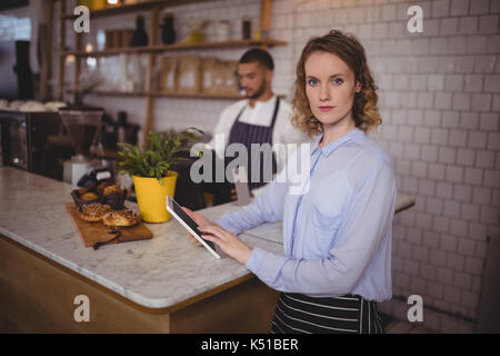 Portrait of smiling waitress using digital tablet while standing at counter in coffee shop Banque D'Images