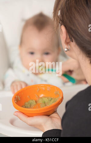 Mère nourrir bébé fille avec la purée d'avocat et de légumes verts dans un bol en plastique orange, de l'alimentation sur une cuillère en plastique ; le concept de la vie familiale, la santé e Banque D'Images
