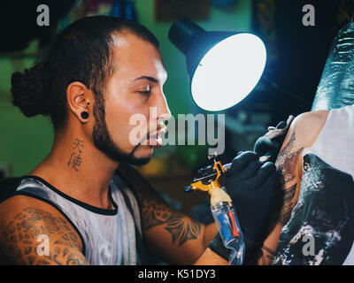 Portrait d'un homme maître de tatouage avec des dreadlocks montrant processus de création tatouage sur le corps féminin sous la lampe de l'artiste travaillant dans sa light.professional Banque D'Images