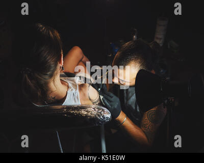 Portrait d'un homme maître de tatouage avec des dreadlocks montrant processus de création tatouage sur le corps féminin sous la lampe de l'artiste travaillant dans sa light.professional Banque D'Images