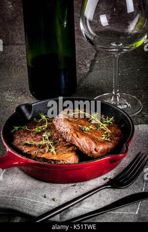 Le déjeuner pour une personne. des steaks de bœuf grillé avec le thym dans une poêle, avec une fourchette, un couteau et un verre de vin sur la table en pierre noire. Banque D'Images