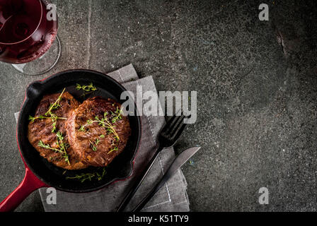 Le déjeuner pour une personne. des steaks de bœuf grillé avec le thym dans une poêle, avec une fourchette, un couteau et un verre de vin sur la table en pierre noire. Banque D'Images