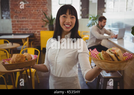 Portrait of waitress holding paniers avec monsieur pendant que businessman using laptop at counter in cafe Banque D'Images