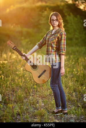 Jolie jeune femme de race blanche en jeans et chemise à carreaux tenir une guitare acoustique à la main sur une prairie d'automne dans l'aube la lumière. Banque D'Images