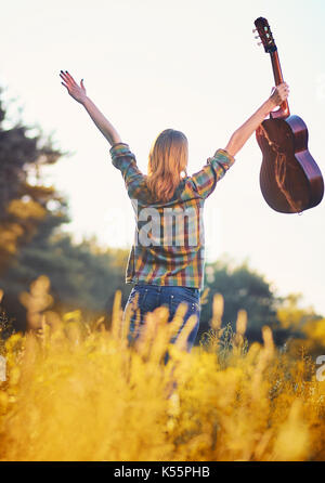 Photo de l'arrière d'un happy girl avec ses mains avec une guitare acoustique sur un pré en automne coucher du soleil la lumière. Banque D'Images