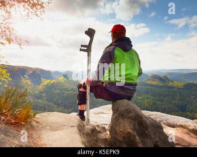 Touriste fatigué avec béquille médecine et jambe cassée fixée à l'antidémarrage reposant sur la montagne de la vallée ci-dessous. l'homme assis dans un coupe-vent vert Banque D'Images