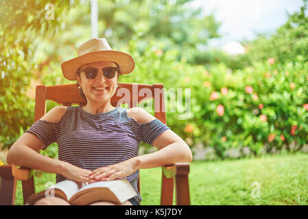 Femme souriante avec livre sur terrasse ensoleillée jardin vert Banque D'Images