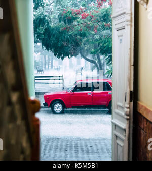 Jour de pluie à Cuba. Vieille voiture rouge aux vitres teintées vu garé sur rue. Banque D'Images