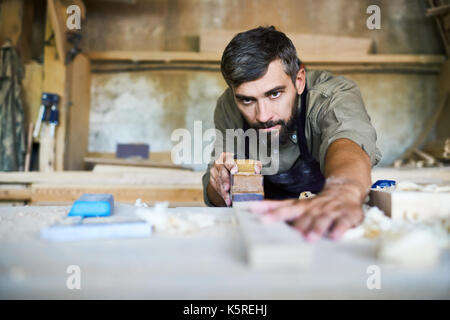 Artisan barbu portant un tablier concentré lissant plank avec plan de la rasette, copeaux éparpillés sur la table Banque D'Images
