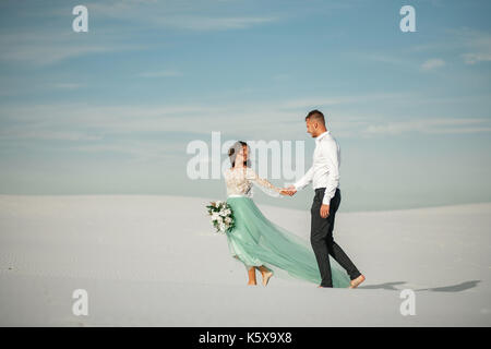 Bride and Groom tenir la main, sourire et marcher pieds nus dans le désert. mariée est habillé en robe de mariage et de bouquet. Ils passe de l'arrière-plan blanc s Banque D'Images