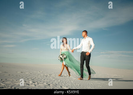 Bride and Groom tenir la main, à rire gaiement et marcher pieds nus dans le désert. mariée est habillé en robe de mariage et de bouquet. Ils passe historique Banque D'Images