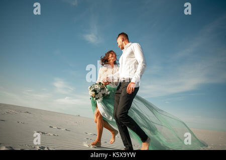 Bride and Groom tenir la main, à rire gaiement et marcher pieds nus dans le désert. mariée est habillé en robe de mariage et de bouquet. Ils passe historique Banque D'Images