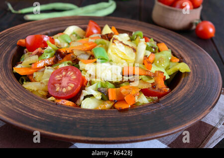 Ragoût aux légumes de choux, haricots verts, carottes, tomates, oignons, poivrons dans un bol d'argile sur un fond de bois foncé. salade végétarienne cu. Banque D'Images