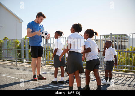 Les jeunes enfants dans une cour d'école avec teacher holding ball Banque D'Images