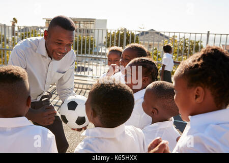 Les jeunes enfants dans une cour d'école avec teacher holding ball Banque D'Images