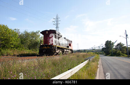 Un train rouge et noir vient le long de pistes en herbe et ciel bleu, derrière la jetée de conteneurs Banque D'Images