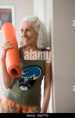 Older woman leaning on wall holding exercise mat Banque D'Images