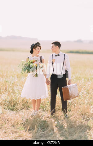 La photo verticale sur toute la longueur de la marche des jeunes mariés habillés vintage et tenir la main dans la zone ensoleillée. l'épouse, c'est maintenant le bouquet de mariage et le marié est portant la vieille valise. Banque D'Images