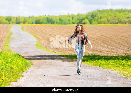 Jeune femme courir, sauter dans l'air et souriant sur la route de campagne par brown champs labourés avec sillons en été à l'île d'Orléans, Québec, Canada Banque D'Images