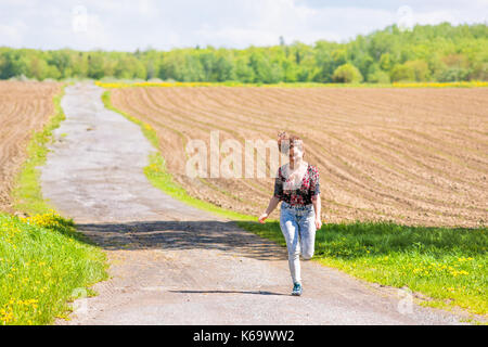 Jeune femme courir, sauter dans l'air et souriant sur la route de campagne par brown champs labourés avec sillons en été à l'île d'Orléans, Québec, Canada Banque D'Images