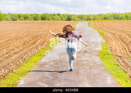 Dos de jeune femme courir, sauter dans l'air et souriant sur la saleté route de campagne par brown champs labourés avec sillons en été à l'île d'Orléans, Québec Banque D'Images