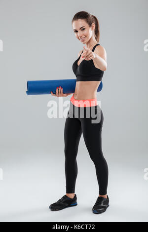 Longueur totale happy woman holding fitness Tapis de fitness et en montrant l'appareil photo sur fond gris Banque D'Images