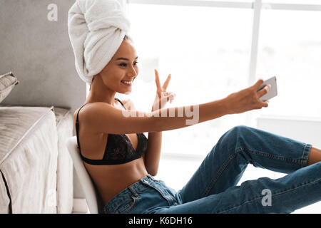 Smiling young asian woman avec bain serviette enroulée autour de sa tête en prenant un tout en selfies assis sur une chaise à la maison Banque D'Images