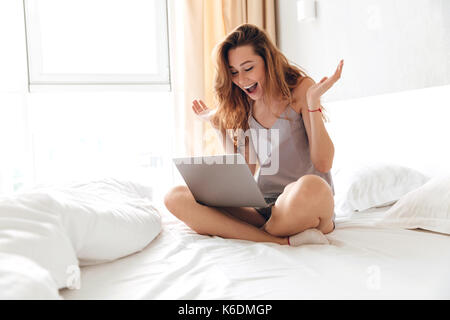 Happy brunette woman using laptop while sitting on lit le matin Banque D'Images