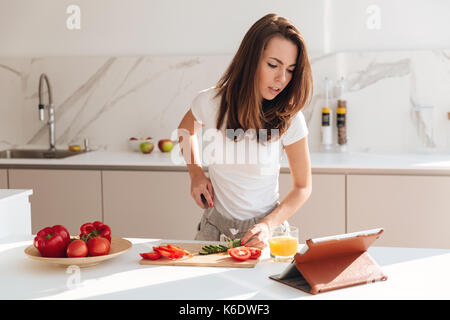 Jeune femme concentré couper des légumes sur une planche en bois et à la tablette à l'ordinateur pendant la cuisson dans une cuisine Banque D'Images