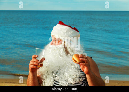 Père Noël sur la plage de manger un hamburger. Le concept de l'alimentation malsaine. Banque D'Images