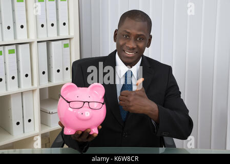 Young happy businessman showing thumb up avec piggybank in office Banque D'Images