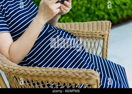 34 semaines de grossesse femme jouant assis smartphone n rotin chaise en bois près du jardin vert Banque D'Images