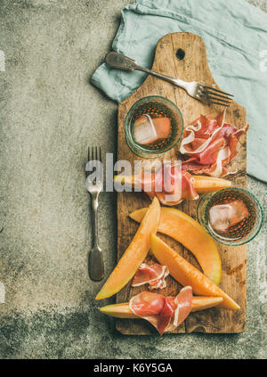 Vin d'été. flatlay partie fixe de vin snack-jambon prosciutto, melon et verres de vin rose avec de la glace sur planche de bois rustique au béton gris background Banque D'Images