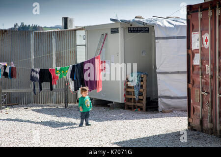 Sur le gravier entre les unités préfabriquées qui hébergent des réfugiés de Syrie, un enfant seul étudie le camp de réfugiés de Ritsona. Foreground : conteneur Croix-Rouge. Banque D'Images