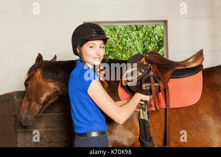 Portrait de la belle jeune femme en uniforme de jockey, debout à l'intérieur de l'école d'un raccord et selle pendant la préparation de son bay horse Banque D'Images