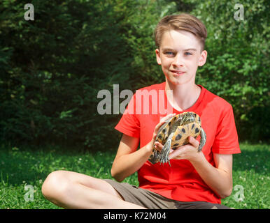 Cute adolescent boy wearing red t-shirt assis sur une pelouse dans un jardin d'été holding turtle looking at camera smiling se concentrer sur turtle Banque D'Images