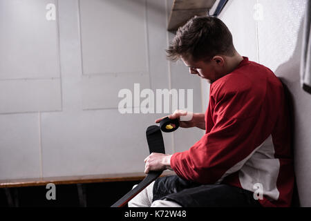 Vue latérale du joueur de hockey sur glace masculin taping stick tout en restant assis dans dressing Banque D'Images